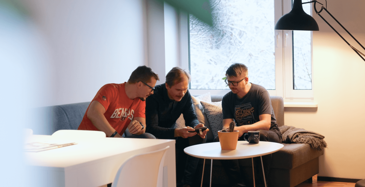 Three men are sitting on a sofa looking at a design view on a phone together, with a table holding coffee mugs in front of them.
