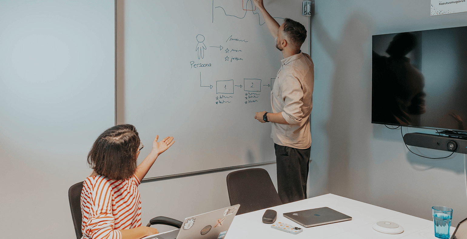 Our two user experience designers: a man drawing a diagram on a whiteboard and a woman at a desk.